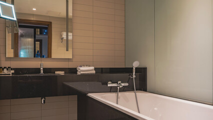 Bathroom interior. A white rectangular bathtub with a shower and taps. On the black marble countertop there is a washbasin, toiletries, rolled towels. A mirror on the tiled wall. Reflection