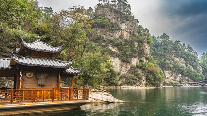 Fototapeta premium The beautiful lake is surrounded by steep picturesque mountains. Green vegetation on the slopes of the cliffs. In the foreground is a Chinese boat with curved roofs and carved wooden railings. China.