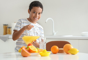 Little African-American boy pouring fresh citrus juice into glass from jug at table in kitchen