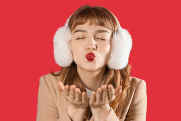 Young woman in earmuffs blowing kiss on red background, closeup