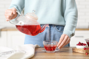 Young woman pouring cranberry tea from teapot into cup in kitchen