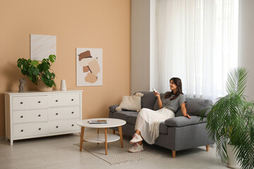 Young woman with air conditioner remote control sitting on sofa near beige wall in living room
