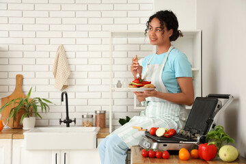 Beautiful young happy African-American woman with plate of vegetables near modern electric grill sitting in kitchen