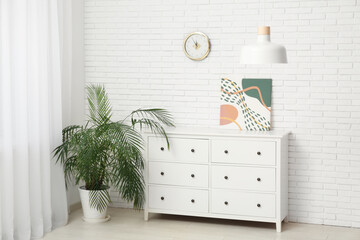 Hanging lamp, chest of drawers with picture and clock near white brick wall in room