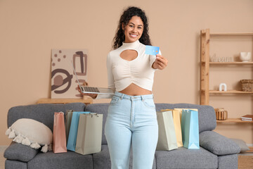Happy young African-American woman with shopping bags, laptop and credit card at home