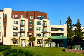 Modern multi-story apartment building with beige and brown exterior, balconies, adjacent to a parking garage, and a manicured lawn in the foreground
