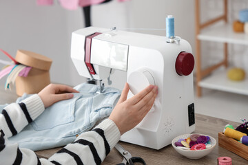 Young female tailor using sewing machine for jeans near table in atelier, closeup