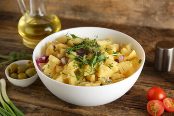 Bowl of tasty potato salad with onion, olives and tomatoes on wooden background
