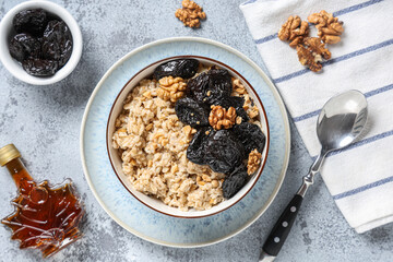 Bowl of tasty oatmeal with prunes, walnuts and bottle of maple syrup on grey grunge background