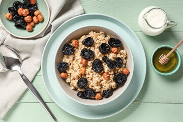 Bowl of tasty oatmeal with prunes, hazelnuts, honey and jug of milk on green wooden background