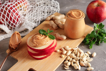 Wooden board with apple rounds, nut butter, peanuts and mint leaves on grey grunge background