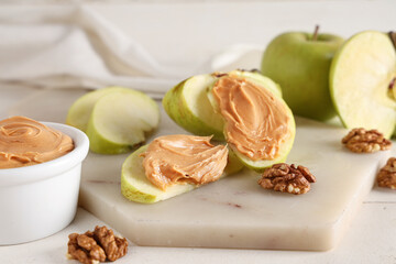 Board with tasty apple wedges, bowl of nut butter and walnuts on white wooden background