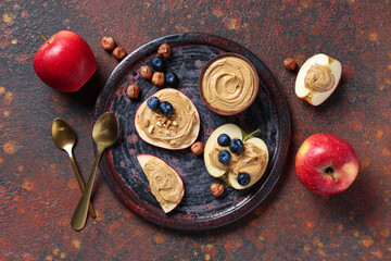 Plate with tasty cut apple, bowl of nut butter, blueberry and hazelnuts on grunge background