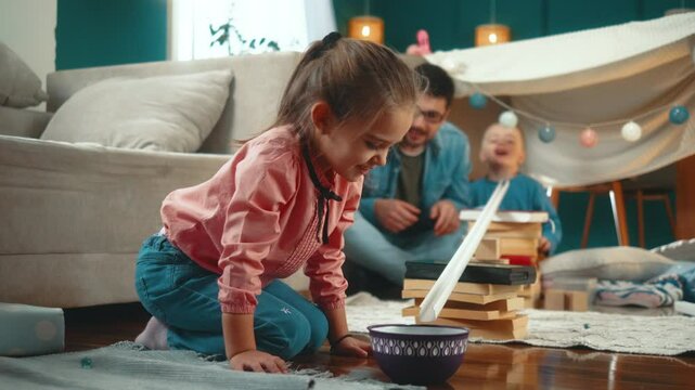 A father and two small children daughter and son play by rolling marbles on a makeshift structure they made in the living room at home