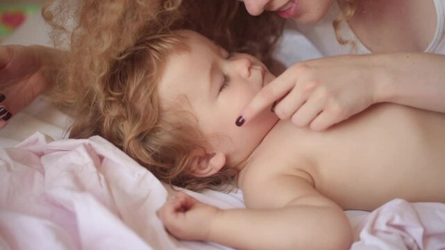 Happy young mother with little baby child lying on bed at bedroom. Beautiful mummy kissing and hug sleeping baby. Mother kissing sleeping baby. Mother caring for lovely child.