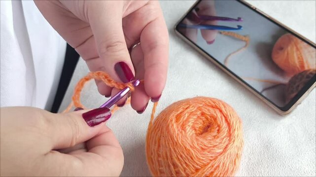 A woman knits a toy based on a video lesson. Online crochet training. A woman's hands are holding a hook and threads, yarn, and in the background is a smartphone with a knitting video.