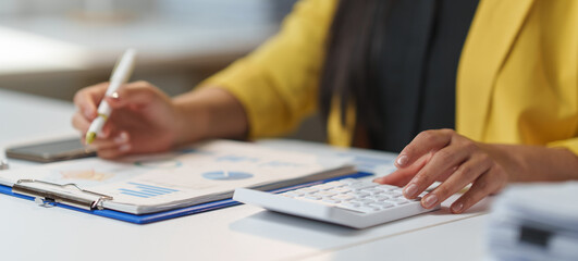 Businesswoman in a yellow blazer analyzing financial reports with a calculator and pen. Focus on data charts and smartphone on a modern office desk