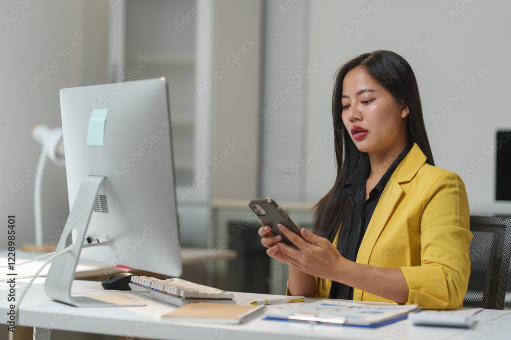 Poster professional woman in a yellow blazer multitasking with a smartphone and computer at a sleek office 