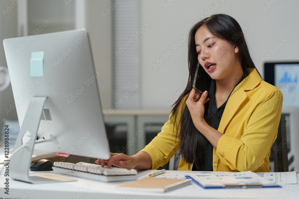 Poster professional woman in a yellow blazer managing phone calls and computer tasks simultaneously at a mo
