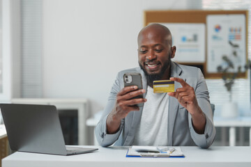 Happy african american manager is holding credit card and using smartphone while sitting at desk in office, making convenient online payment