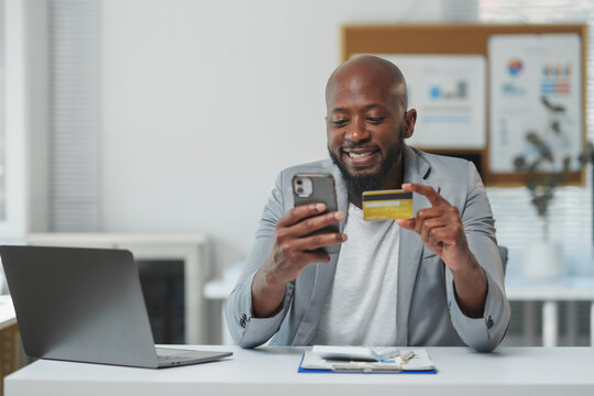 Happy african american manager is holding credit card and using smartphone while sitting at desk in office, making convenient online payment