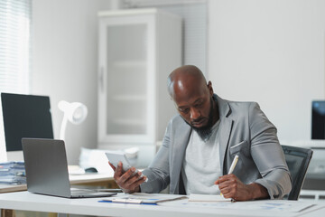 Concentrated african american male accountant in gray suit sitting at desk in office, using...