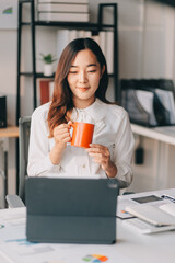 Young asian businesswoman sitting on an ergonomic chair and drinking coffee while working on a laptop computer in a modern office, she is smiling and looking at the screen