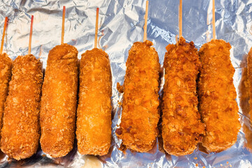 Fried battered corn dog on wooden sticks on a foil plate, asian street food. Sausage and cheese wrapped in flour batter. Close-up of roasted crunchy snack on skewer coated crispy bread crumbs.