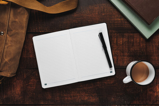 Open notebook with a pen on a rustic wooden surface, accompanied by a canvas bag, books, and a coffee mug, creating a cozy and productive workspace scene.