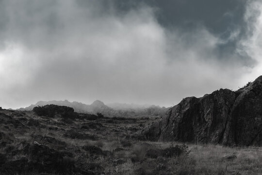 Dark, rocky landscape under a heavy, cloudy sky. Black and white photography of a gloomy and dramatic atmosphere.  Rugged terrain with sparse vegetation.