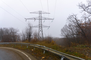 High voltage power pole on the edge of the asphalt road. Iron barrier and white stripe on the edge of the road. Bushes and thick fog in the background