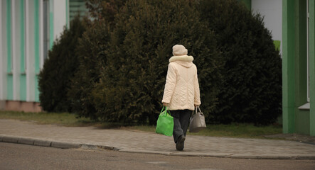 an elderly woman in a light jacket walks with heavy bags down the street