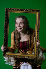 Obraz premium Woman in ancient outfit on chroma key green screen background. Female in renaissance dress holding antique frame and posing as portrait painting at camera.