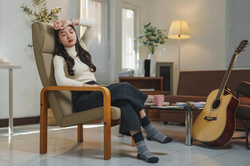 Fototapeta premium Young asian woman sitting in a cozy armchair with socks on, resting her head in her hand, feeling exhausted and stressed, with an acoustic guitar nearby