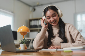 Young asian woman working from home office, wearing headphones, listening to music and taking notes, with laptop and desk lamp in background