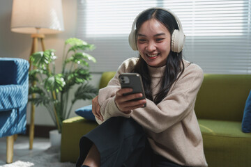Smiling young woman with headphones enjoying music while looking at her smartphone. Sitting on a comfortable couch in a cozy living room with natural light and indoor plants