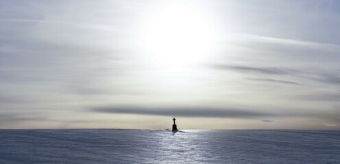 Fototapeta premium A lighthouse stands alone in the sea under a wide sky while the sun sets in the background, a lonely field cross in the snow