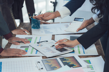 Strategic Collaboration: Hands of a diverse business team huddle around a table strewn with financial reports, charts, and graphs, collaboratively analyzing data and strategizing.  