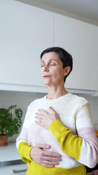Peaceful mature woman engages in deep breathing exercises with hands placed on chest at home. Relaxed female mindfulness, serenity and stress free meditation, slow time with yourself in solitude