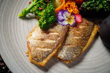 Pan-seared fish fillet with vibrant vegetables, including broccoli, cherry tomatoes, and carrot ribbons, garnished with edible flowers. Accompanied by a dark sauce jug. Fine dining closeup