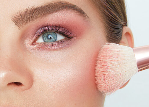 Close up beauty portrait of a woman applying pink blush with a fluffy makeup brush. Her blue eye is highlighted with shimmering eyeshadow, and her eyebrows are well-groomed.