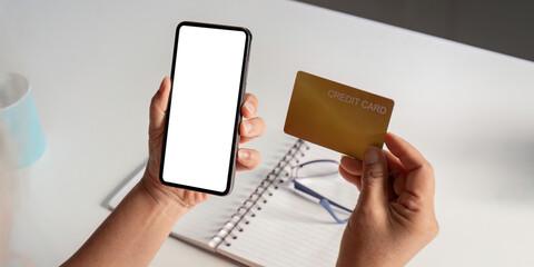 Elderly person holding a smartphone with a blank screen and a credit card over a table.