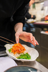A chef skillfully garnishes a dish with salmon and fresh greens using chopsticks in a bustling...