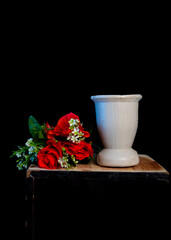 Fresh red roses with green leaves and white blossoms beside an empty white vase, set on a wooden surface against a black background.