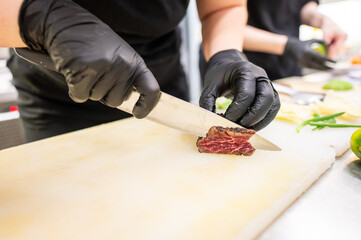 A chef in black gloves skillfully slices a piece of seared meat on a cutting board in a professional kitchen, showcasing culinary precision and attention to detail.