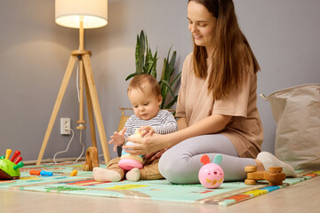 Fototapeta premium A mother sits on the floor, playfully interacting with her infant. They are surrounded by colorful toys on a soft play mat in a bright, welcoming living room space