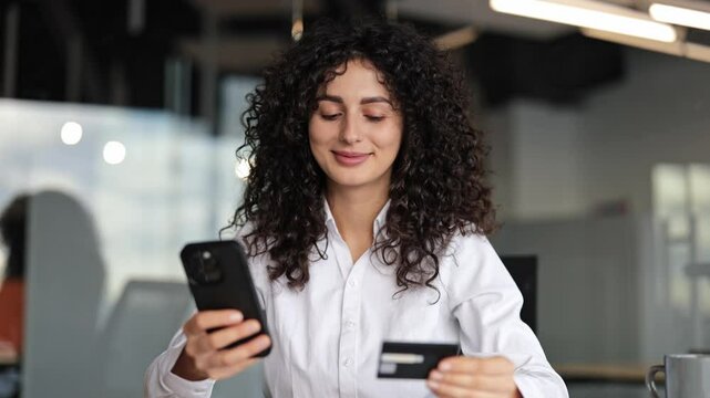 Confident young businesswoman using smart phone and credit card for online shopping in modern office. Curly haired female employee making secure payment transaction via mobile app.