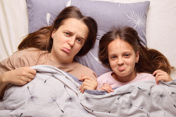 A mother and her daughter are lying in bed, both showing exaggerated, pouty expressions. They are...
