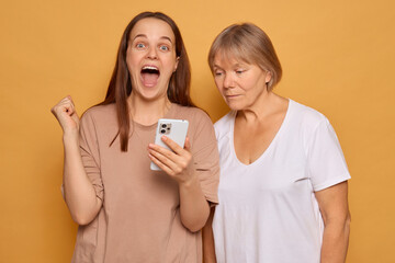 Two women express contrasting emotions while looking at a smartphone. One shows joy with an...