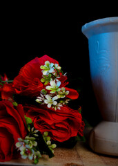 Fresh red roses with green leaves and white blossoms beside an empty white vase, set on a wooden...